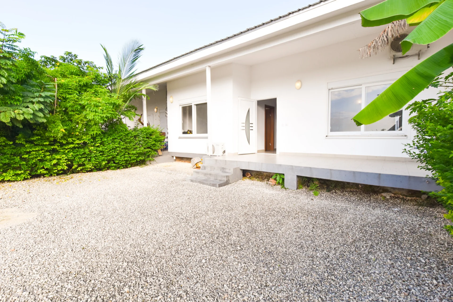 White single-storey townhouse facing a gravel garden courtyard edged with tropical greenery.